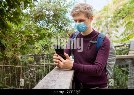 Junger Mann mit Gesichtsmaske, Filmen von Videos oder Fotografieren am Telefon, während er während der Coronavirus-Pandemie in einem exotischen Park unterwegs ist. Neue norm Stockfoto