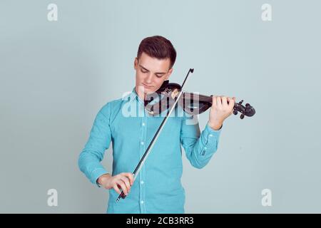 Musikleidenschaft, Hobby-Konzept. Der junge Mann, der modern und elegant gekleidet ist, spielt auf einer Holzvioline in einem Studio, das auf einem hellblauen Wandhintergrund isoliert ist. Ho Stockfoto