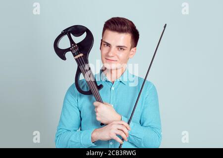 Junger Mann posiert halten moderne Geige und Bogen lächelnd über Arme im Studio isoliert hellblau Wand Hintergrund. Horizontales Schussmodell in blauen Cla Stockfoto