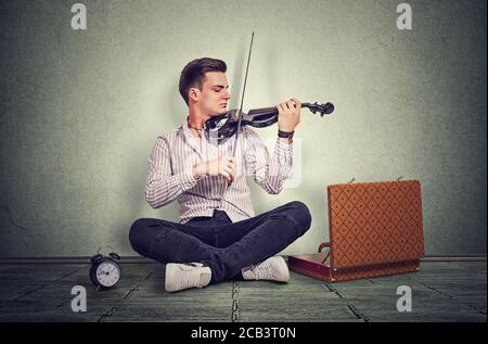 Der junge Mann, der modern gekleidet ist, spielt auf einer schwarzen elektronischen Geige in einem Studio, das auf grauem Boden vor einem grauen Wandhintergrund sitzt. Es ist Zeit zu machen Stockfoto