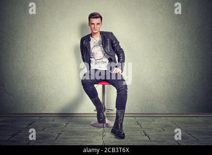 Portrait von selbstbewussten jungen Mann auf einem roten Stuhl isoliert auf grauem Hintergrund sitzen, Boden im Studio isoliert auf grauem Wandhintergrund. Horizontale Aufnahme Stockfoto