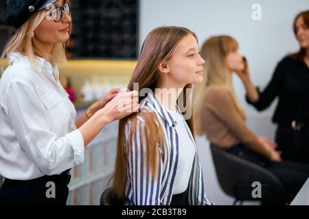 Vorsichtig kaukasischen Friseur hält Client Haar in den Händen, gehen, um Spitzen der Haare schneiden, so dass Frisur für junge Mädchen Client sitzt auf Stuhl oppos Stockfoto