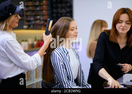 Professionelle moderne Friseur glätten Haar junge schöne Frau auf Stuhl sitzen, Blick auf Spiegel während des Prozesses der Herstellung von Frisur in Schönheit sa Stockfoto