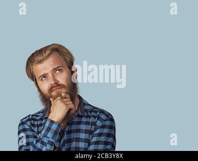 Verwirrter skeptischer Mann, der denkt, mit dem Kopierraum über dem Kopf nach oben zu schauen. Hipster Männchen mit Bart in blau kariert kariertes Hemd isoliert auf blauem Studio Stockfoto