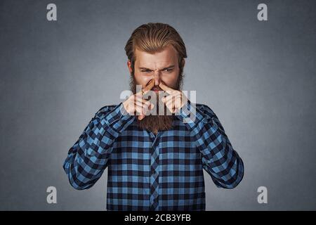 Mann kneift Nase mit Fingern sieht mit Ekel etwas stinkt schlechten Geruch. Hipster Männchen mit Bart in blau kariert kariertes Hemd isoliert auf grau g Stockfoto