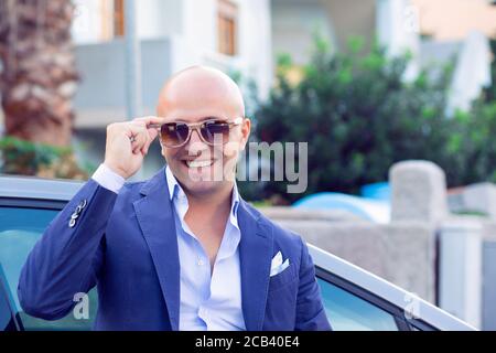 Glücklicher Geschäftsmann, der Sie lächelnd anschaut, aufgeregt hält, justierend Sonnenbrille in blau marineblauem Anzug und weißem Knopfhemd Auf Stadt städtischen Backgr Stockfoto