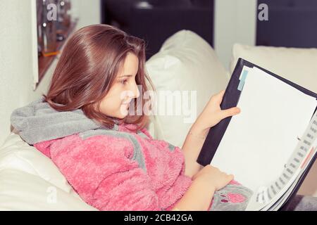 Seitenansicht der jungen legeren Frau im Pullover sitzend auf Sofa mit Notizblock und Lesestoff zu Hause Stockfoto
