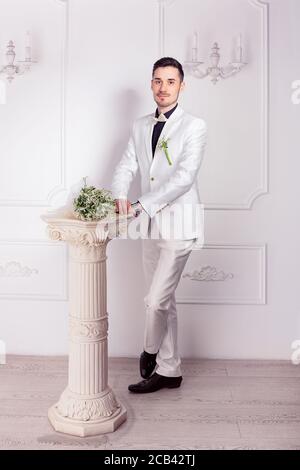 Trendy Bräutigam in weißem Anzug und schwarzen Schuhen stehen mit brautstrauß auf kleiner Säule in der Nähe Blick auf die Kamera Stockfoto
