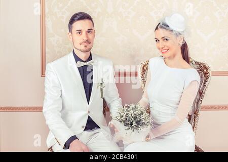 Mann und Frau in eleganten Hochzeitsoutfits sitzen auf Stühlen Und die Hände halten, um die Kamera zu betrachten Stockfoto