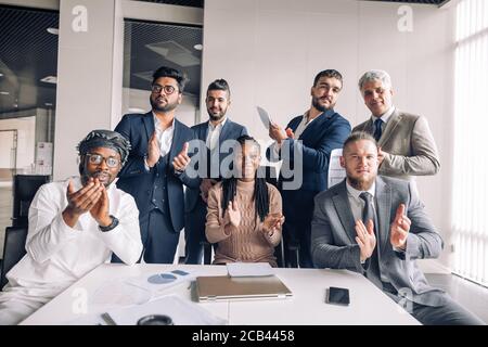 Zufrieden zufrieden Business Männer verschiedener Rasse und Alter, gekleidet in formale zusammen mit jungen afrikanischen Frau Applaus an die Kamera nach Visitin Stockfoto