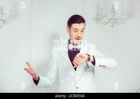 Zu spät. Junger emotionaler Mann im trendigen Bräutigam Anzug, der Armbanduhr mit Überraschung auf weißem Hintergrund anschaut. Sie ist zu spät für den Eheprozess con Stockfoto