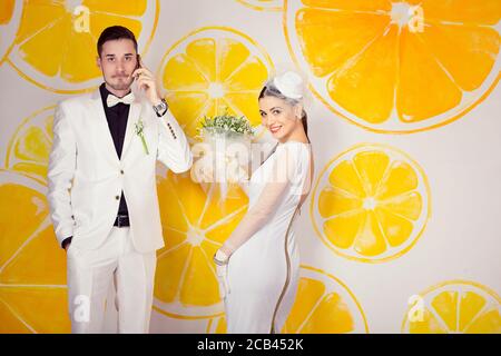 Trendy fröhlich Braut im Kleid posiert Holding Bouquet während Bräutigam Telefoniert mit Orangen in der Nähe der Wand Stockfoto