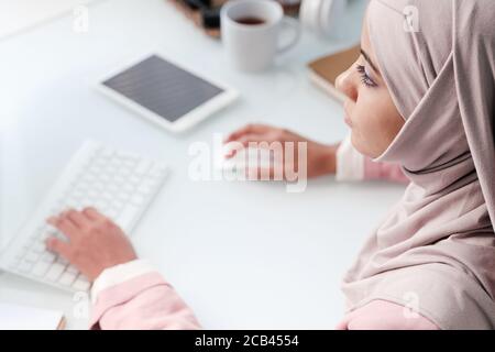 Fokussierte junge muslimische Geschäftsfrau in traditionellen Hijab sitzen am Tisch Und mit dem Computer Stockfoto