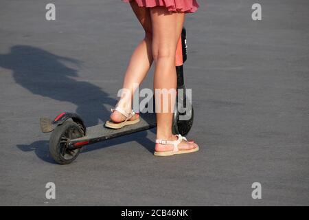 Mädchen reitet einen Elektroroller auf einer Stadtstraße, schlanke weibliche Beine auf Asphalt. Reiten E-Scooter im Sommer Stockfoto