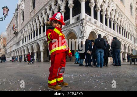 VENEDIG, ITALIEN - 28. JANUAR 2018: Ein Kind mit Feuerwehrkostüm, während des Karnevals von Venedig mit dem Markusplatz im Hintergrund Stockfoto