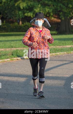 An einem Sommermorgen während der Pandemie, läuft eine Frau trägt eine chirurgische Maske Gesichtsschutz & helle Jacke. In Kissena Park, Flushing, New York City. Stockfoto