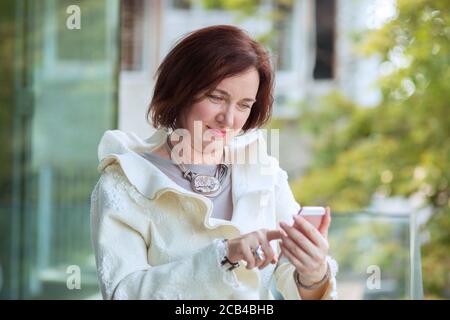 Moderne reife Brünette ruht auf der Terrasse und Surfen Handy Stockfoto