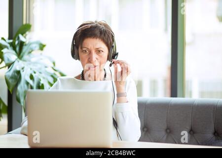 Moderne reife Frau im Headset mit Videoanruf mit Laptop Beim Sitzen am Tisch im Café Stockfoto
