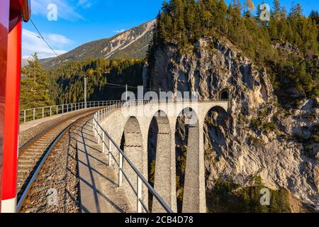 Fahren Sie mit der roten Rhätischen Eisenbahn-Sightseeing-Zug Bernina Express über Landwasser Viadukt an sonnigen Herbsttag mit blauen Himmel Wolke, Canon Stockfoto