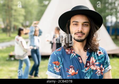 Porträt des Inhalts schöner jüdischer Kerl in schwarzem Hut und shirt mit Blumendruck, der mit Freunden auf dem Waldcampingplatz ruht Stockfoto