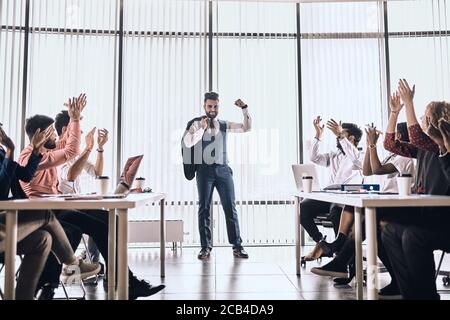 Teamleiter zeigt seinen Bizeps, während Kollegen ihm applaudieren. Stärke der Firma. Chef motiviert seine Kollegen. Motivation Stockfoto