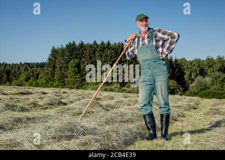 Freundlicher, lächelnder ökologischer Bauer steht auf seinem Bio-Heufeld in der Sonne und macht eine Pause von der schweißtreibenden harten Arbeit. Stockfoto