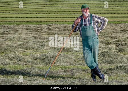 Freundlicher, lächelnder ökologischer Bauer steht auf seinem Bio-Heufeld in der Sonne und macht eine Pause von der schweißtreibenden harten Arbeit. Stockfoto