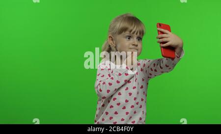 Kind Mädchen mit Smartphone. Porträt des Kindes mit blonden Haaren macht emotional Selfie auf Handy. Videoanruf, Blog, Spiele spielen, Handy ansehen. Mit lustigen Gesichtern. Chroma-Taste Hintergrund Stockfoto