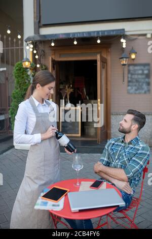 Fröhliche Kellnerin, die dem bärtigen Mann eine Weinflasche zeigt und im Open-Air-Café sitzt Stockfoto