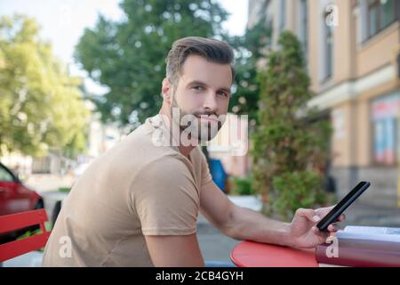 Bärtiger Mann sitzt im Café, hält Smartphone, lächelnd Stockfoto