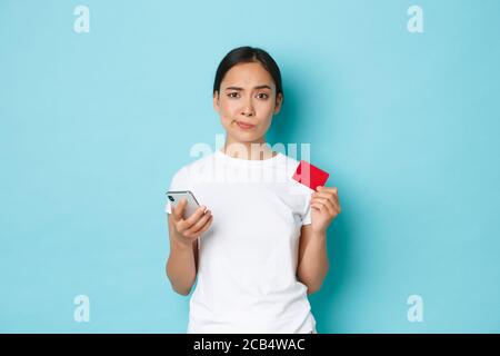 E-Commerce, Shopping und Lifestyle Konzept. Skeptisch und zweifelhaft asiatische Mädchen suchen unzufrieden, grinsen, während halten Smartphone und Kreditkarte Stockfoto