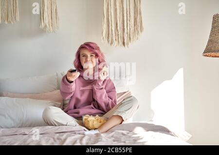 Lächelndes Hipster Gen Z Teenager-Mädchen mit rosa Haaren Fernsehen essen Chips im Bett. Stockfoto