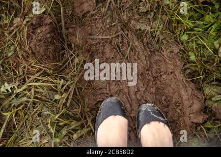 Zwei Füße tragen schwarze Ballet flache Schuhe mit Schlamm bedeckt stehen auf dem schlammigen Druck eines LKW zwischen dem grünen Gras. Stockfoto