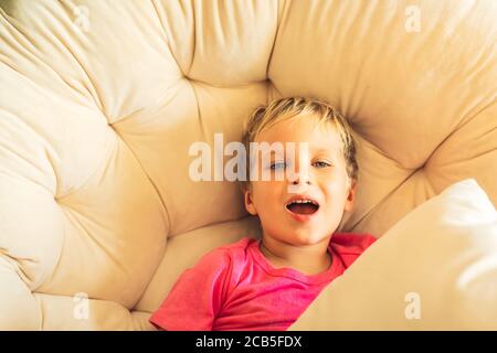 Blond lachend mit Spaß junge offenen Mund in rosa T-Shirt sitzen mit Kissen in runden Stuhl innen. Familienbeziehung, glückliche Kindheit, Problembehaviou Stockfoto