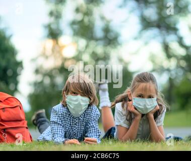 Die Kinder gehen zurück zur Schule. Nette Schüler mit Rucksäcken. Junge und Mädchen in Sicherheitsmasken liegen auf dem Gras. Stockfoto