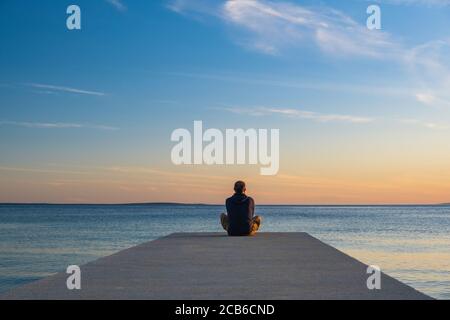 Mann, der am Rande des Docks sitzt und meditiert. Meereshorizont bei Sonnenuntergang, Insel von Pag, Adria, Kroatien. Stockfoto