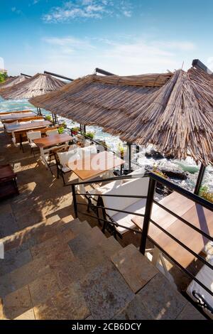 Leere Terrasse eines Restaurants am Meer. Schöner Blick in die Bucht im Morgenlicht. Dekorativer Schirm über Holztischen und Sitzplätzen. Sonniges Wetter Stockfoto