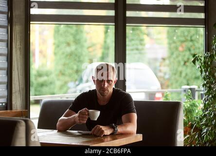 Portrait des sportlichen Mannes ist mit einer Tasse Kaffee in einem Café, Blick auf die Kamera. Riesige Fenster mit seinem Auto hinter ihm auf dem verschwommenen Hintergrund Stockfoto