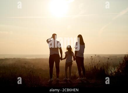 Silhouette einer Familie - Vater, Mutter und Tochter stehen auf dem Hügel mit dem Rücken zur Kamera und blicken zum Horizont bei Sonnenuntergang Stockfoto