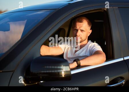 Selbstbewusster ernster Mann mit Borsten in weißem T-Shirt sitzt in seinem schwarzen Auto. Lächelnder Mann, der aus dem Fenster auf die Kamera schaut, Nahaufnahme Stockfoto