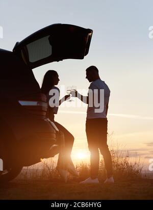 Vertikale Aufnahme Silhouette eines Paares, Mann und Frau, mit romantischen Zeit trinken Wein im Kofferraum auf dem Hügel auf dem Sonnenuntergang, Abendhimmel auf dem Hintergrund, kopieren Raum Stockfoto