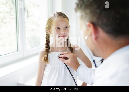 Männlicher Arzt im weißen Mantel untersucht junge, hübsche Mädchen in seiner Praxis Stockfoto