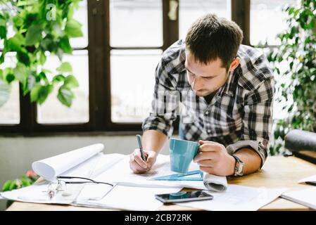 Architekt, der an einem freiberuflichen Renovierungsprojekt in seinem coolen Home-Office-Studio arbeitet. Junger Freiberufler zeichnet seine kreative Design-Idee für Remote-Job. Stockfoto