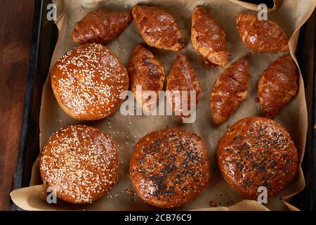 Gebackene Croissants und runde Brötchen mit Sesamsamen auf einem Eisenbackblech mit gerolltem braunem Backpapier, von oben betrachtet Stockfoto