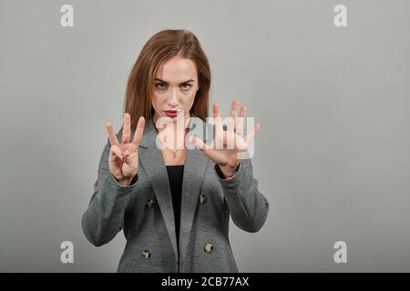 Zeigt acht, 8 Finger Handbewegung, zeigen Sie die Zahl drei mit den Händen, zeigt nach oben Arm, während lächelnd zuversichtlich, glücklich. Junge attraktive Frau, gekleidet graue Jacke, mit grünen Augen, hellbraune Haare Stockfoto