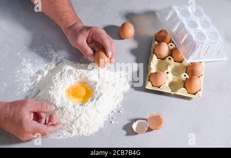 Männliche Hände greifen ein Ei über einem Mehlhaufen umgeben von Eierschalen und einem Karton Eier auf einem Tisch. Backkonzept. Stockfoto