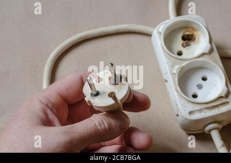 Ein Elektriker demonstriert einen verbrannten Stecker. Weißes Verlängerungskabel nach Kurzschluss in der männlichen Hand. Elektrische Sicherheit. Stockfoto