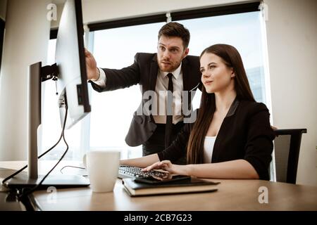Krisenkonzept. Junge Geschäftsfrau sitzt am Bürotisch beschäftigt am Computer arbeiten. Geschäftsmann zeigt auf Monitor. Lösung eines ernsten Stockfoto
