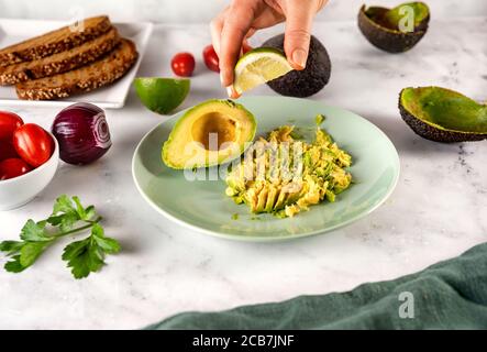 Die Hand der Frau drückt eine Scheibe Limette auf eine pürierte Avocado In einer Platte Stockfoto