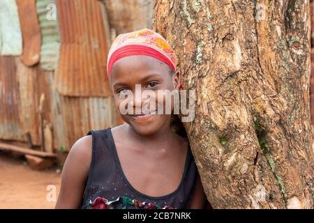 Ein schönes Mädchen im Teenageralter lächelt und lehnt sich an einen Baum. 2017 in Uganda gedreht. Stockfoto